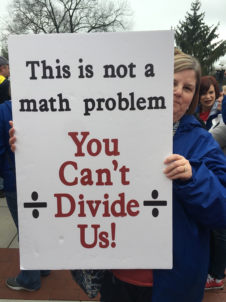 [Photo: A protester holds a sign that reads "This is not a math problem. You can't divide us!]