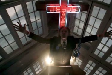 [PHOTO: A man in a three-piece suit stretches his arms skyward, while he stands before a lectern, a red-neon "Jesus Saves" sign, and tall windows]