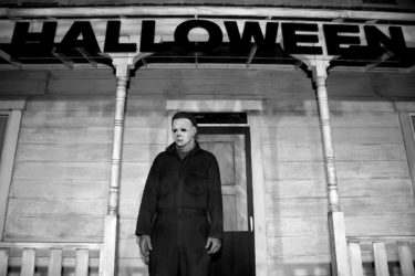[PHOTO: Man in white face mask and black clothing stands in front of a white clapboard house with the words "Halloween" dangling from its eaves.]
