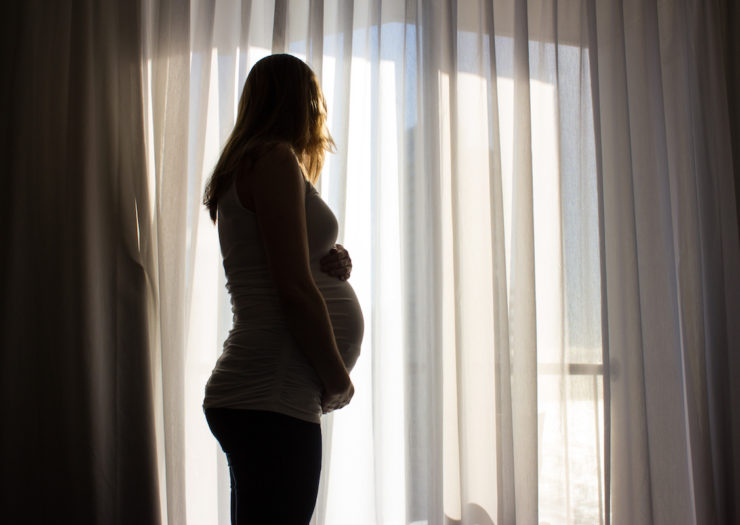 [Photo: Silhouette of a pregnant woman holding her belly looking out a window.]