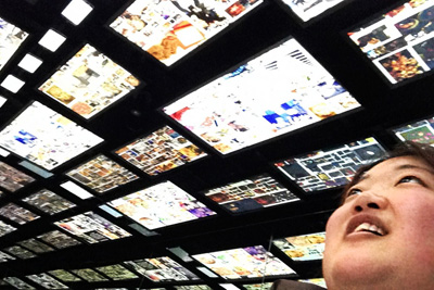 [Photo: A woman is positioned in the corner, looking up at a ceiling full of TV screens.]