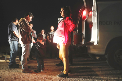 [Photo: A pregnant migrant and others stand next to a United States Border Patrol truck during the night.]